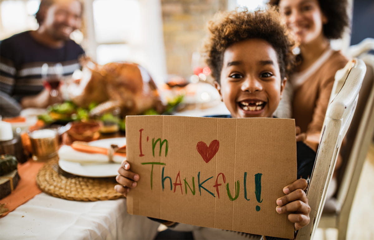 A kid is smiling and holding a sign "I'm Thankful" while seating at the holiday table